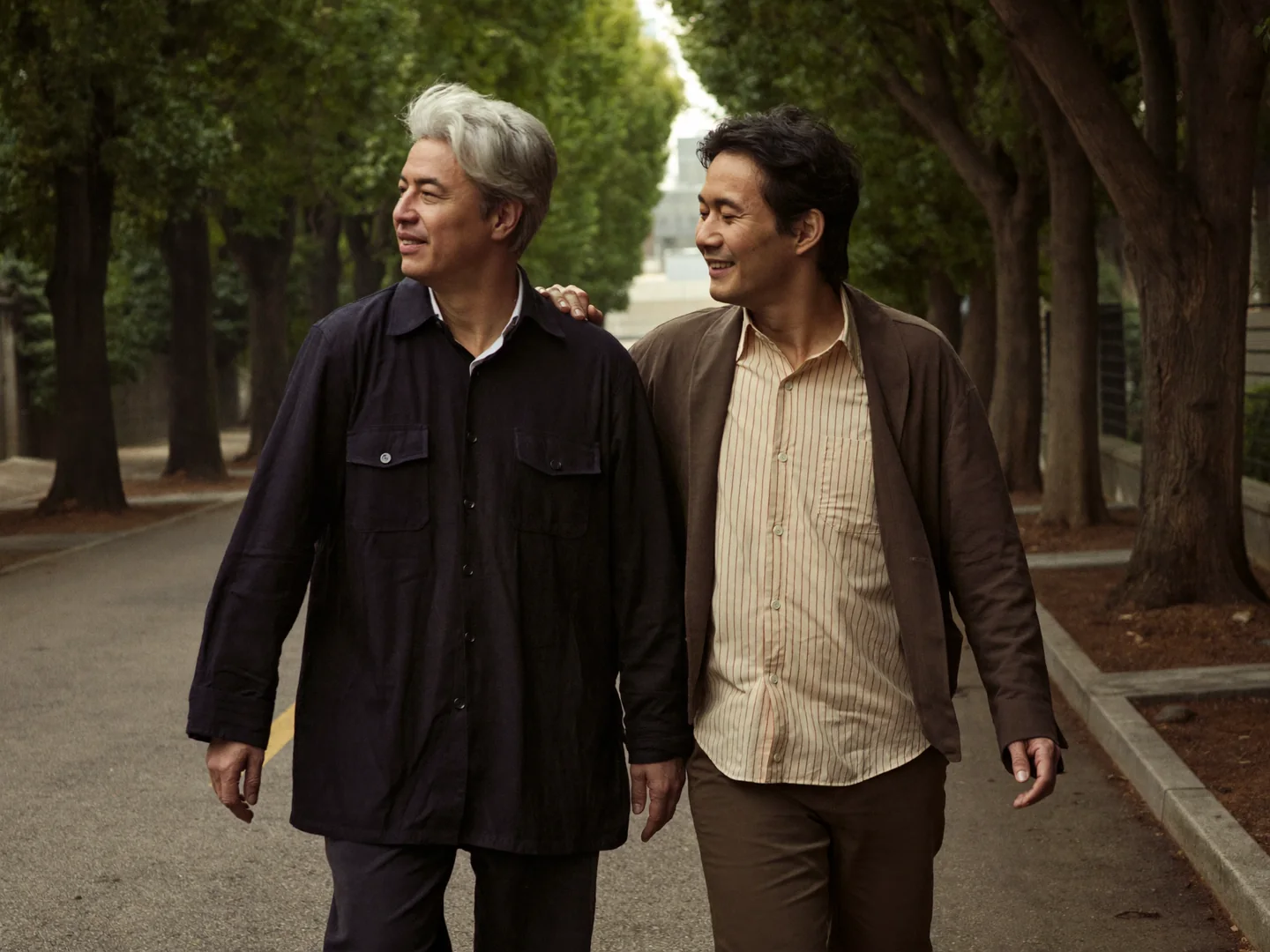 Two men walking closely together along a leafy street.